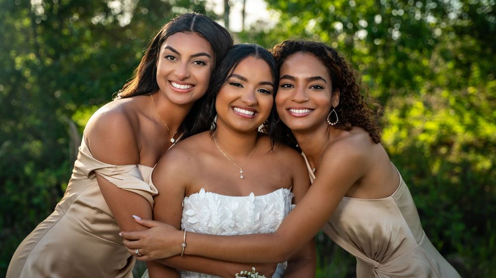 Bride in white strapless dress smiling with two bridesmaids in champagne gowns outdoors.