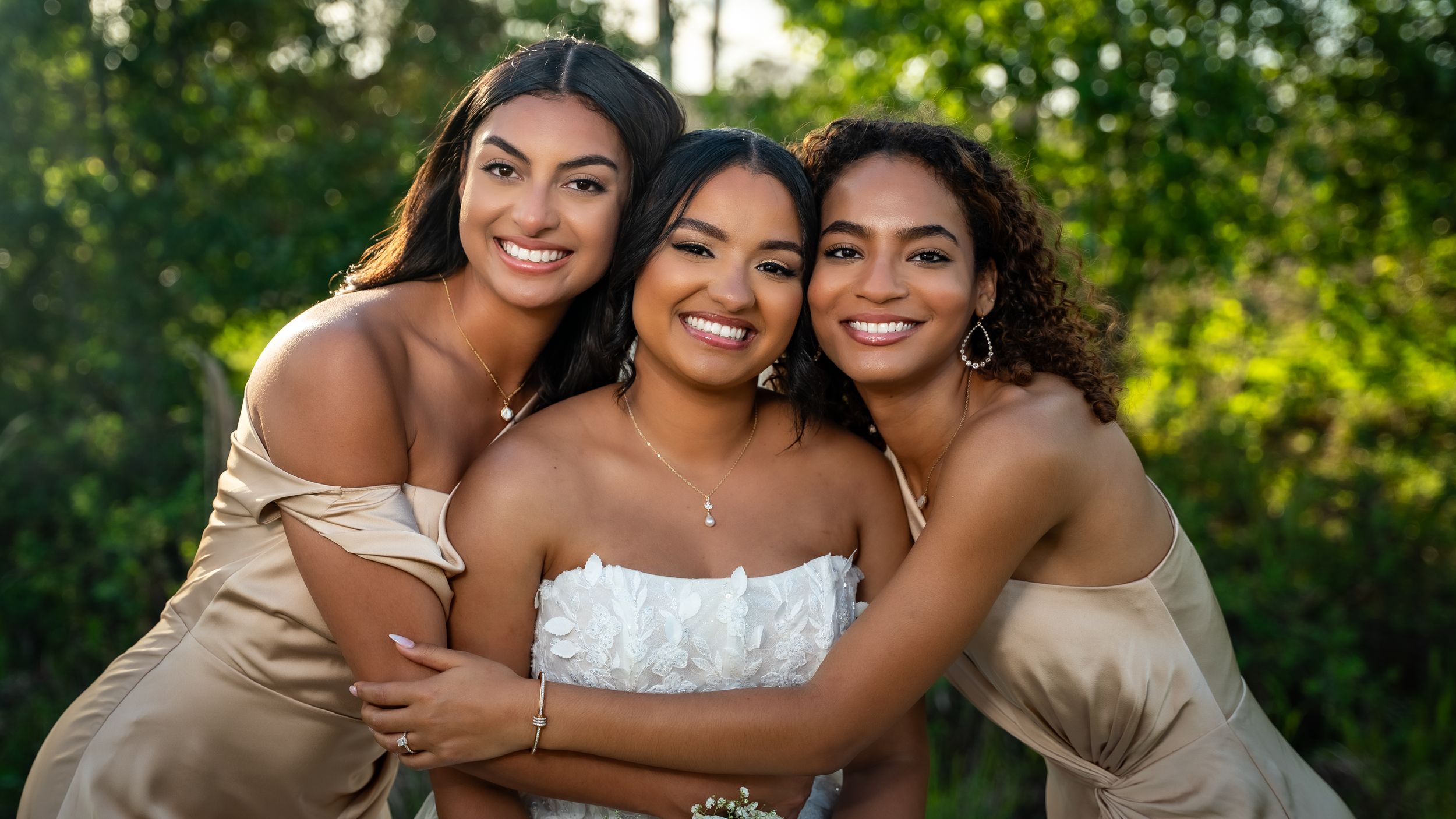 Bride in white strapless dress smiling with two bridesmaids in champagne gowns outdoors.