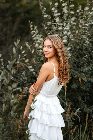 Woman in white ruffled dress poses in garden setting with natural greenery.