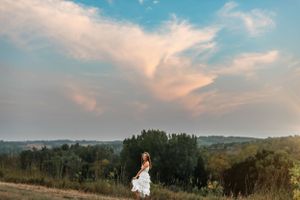 Woman in white dress stands against dramatic sky at sunset on hilltop.
