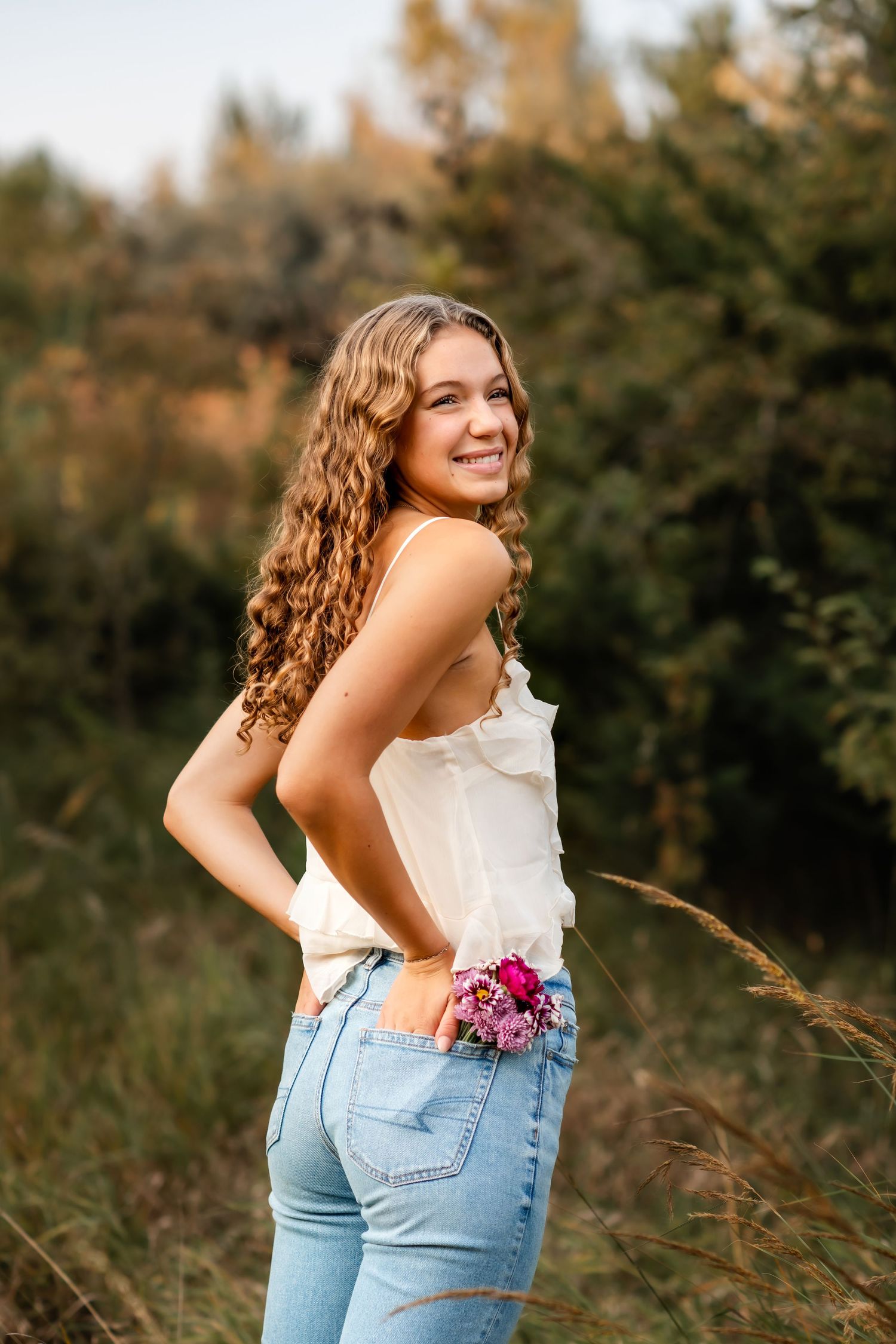 Young woman in white top and jeans poses outdoors with flowers at sunset.
