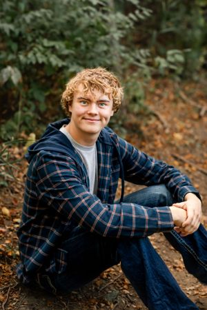 Person in plaid shirt sitting casually on forest path surrounded by greenery.