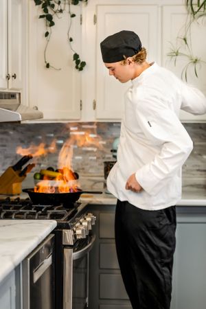 Chef in white uniform working at stovetop with flaming pan in modern kitchen.