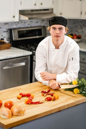 Chef in white uniform prepping ingredients on wooden cutting board in professional kitchen.