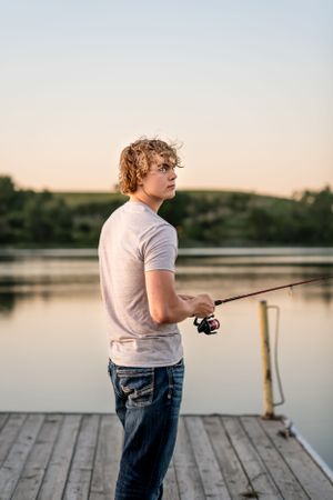 Person in white t-shirt fishing from wooden dock at dusk.