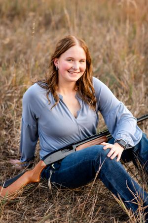 Woman in blue top holding hunting rifle while sitting in tall grass field.