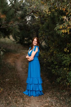 Woman in flowing blue tiered maxi dress poses on wooded trail during autumn.