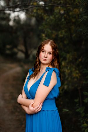 Portrait of a woman in a blue dress with bow details standing against a dark natural background.