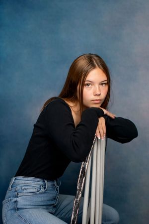 Senior in black sweater and jeans poses against blue background while leaning on a chair for a senior studio session.
