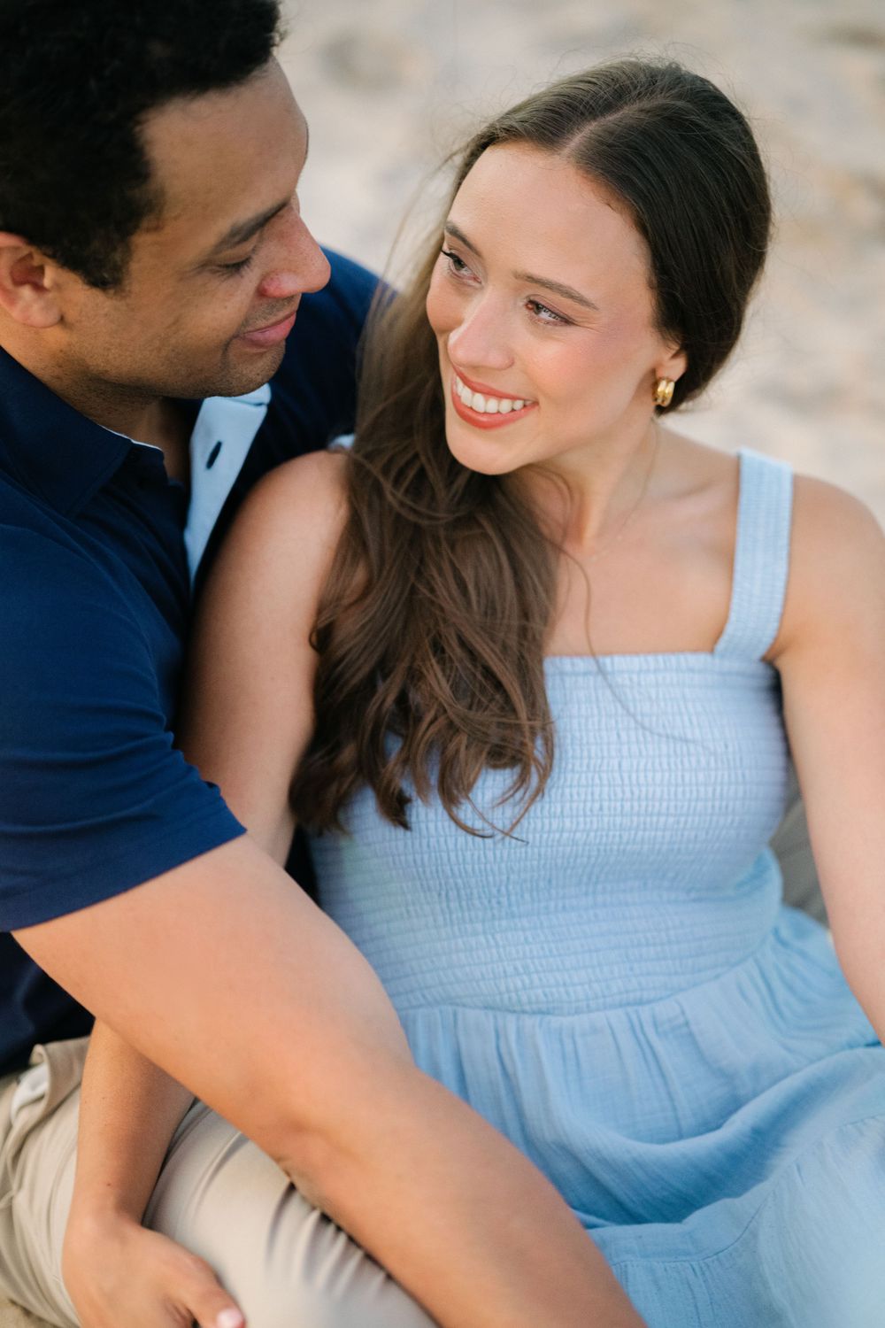 A smiling woman in a light blue dress sits closely with a man in a navy polo on a sandy beach. - North Avenue Beach