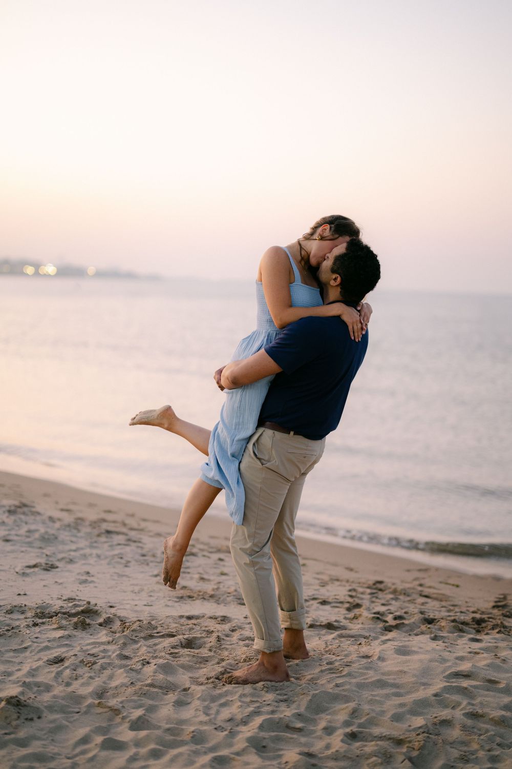 Couple sharing a romantic kiss on a sandy beach at sunset, man lifting woman in a blue dress by the ocean.