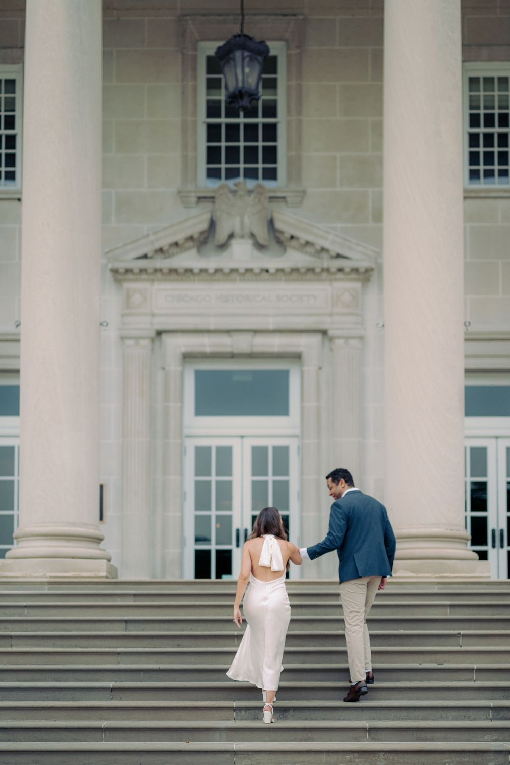 Couple walking up grand stone steps of a neoclassical building, woman in white dress, man in blue suit. - Chicago History Museum Engagement