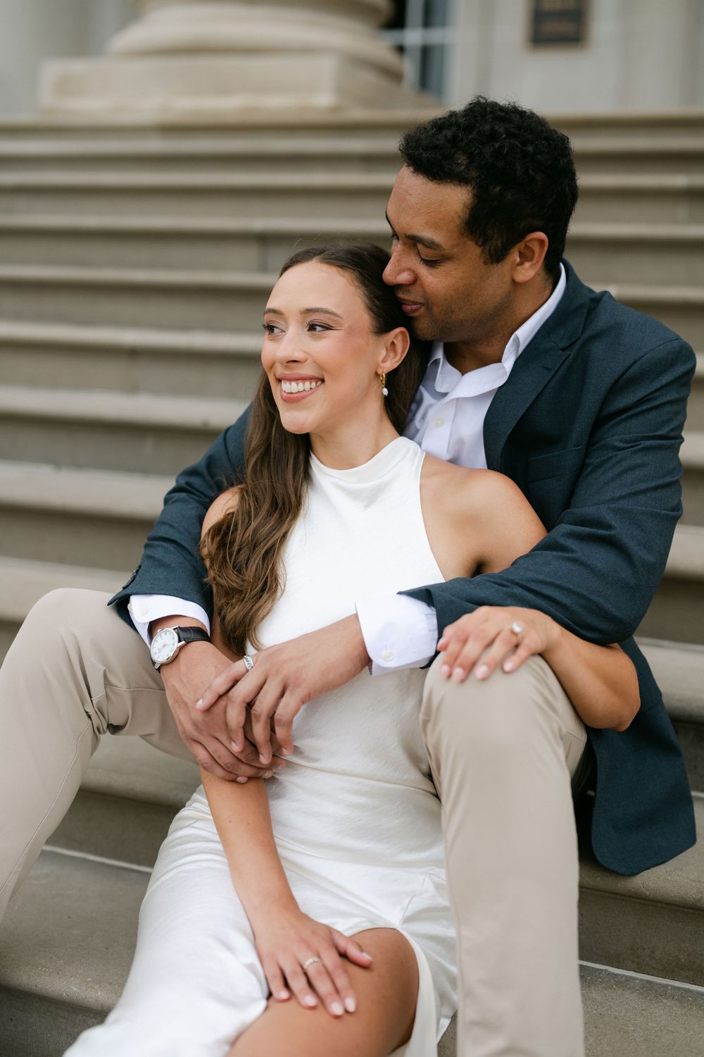 A smiling couple sits on stone steps, woman in white dress, man in navy blazer embracing her tenderly.
