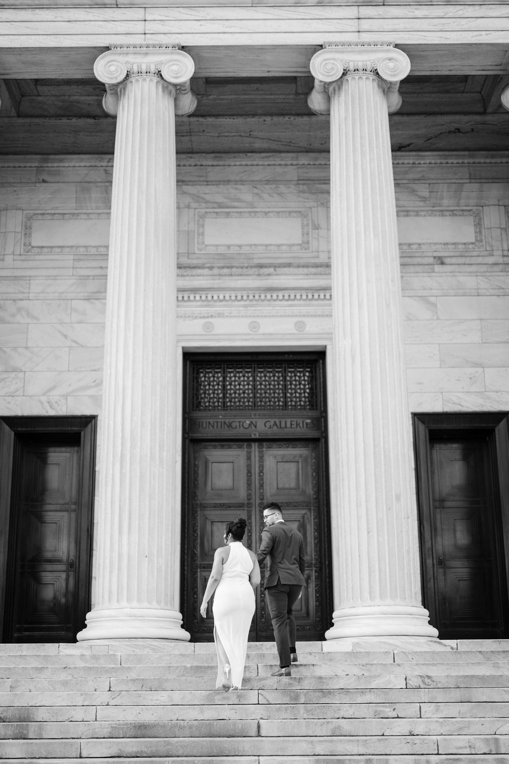 Couple ascending stone steps between grand Ionic columns at entrance of neoclassical building in black and white.