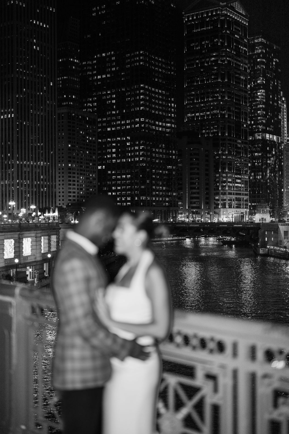 Engagement - Black and white night photo of a couple embracing on a bridge with Chicago River and illuminated skyscrapers in background.