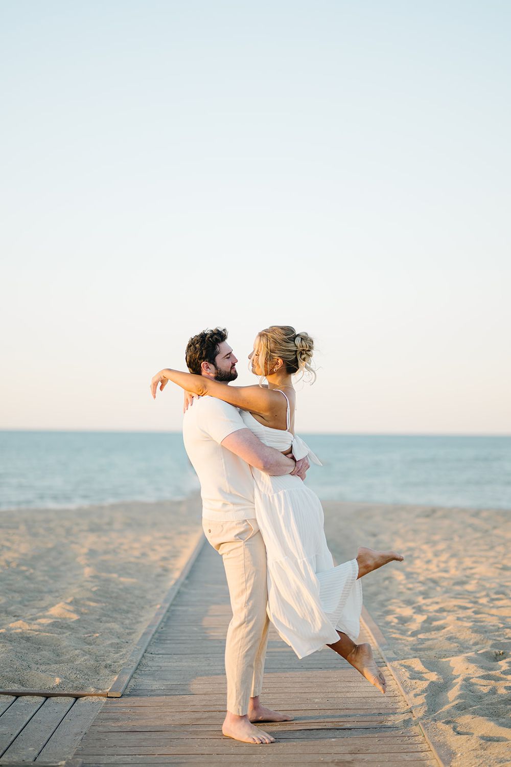 A couple embraces on a beach boardwalk at sunset, the man lifting the woman in a white dress near the ocean.  - North Ave Beach Engagement