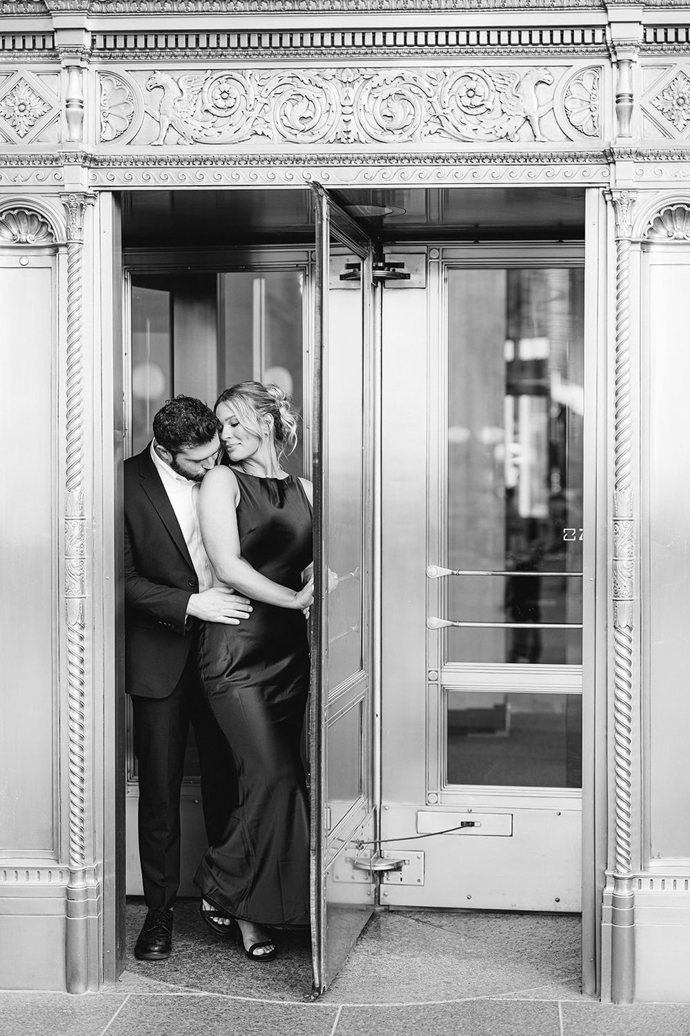 Black and white photo of a couple embracing in an ornate revolving door, dressed in elegant formal attire. - Wrigley Building Engagement