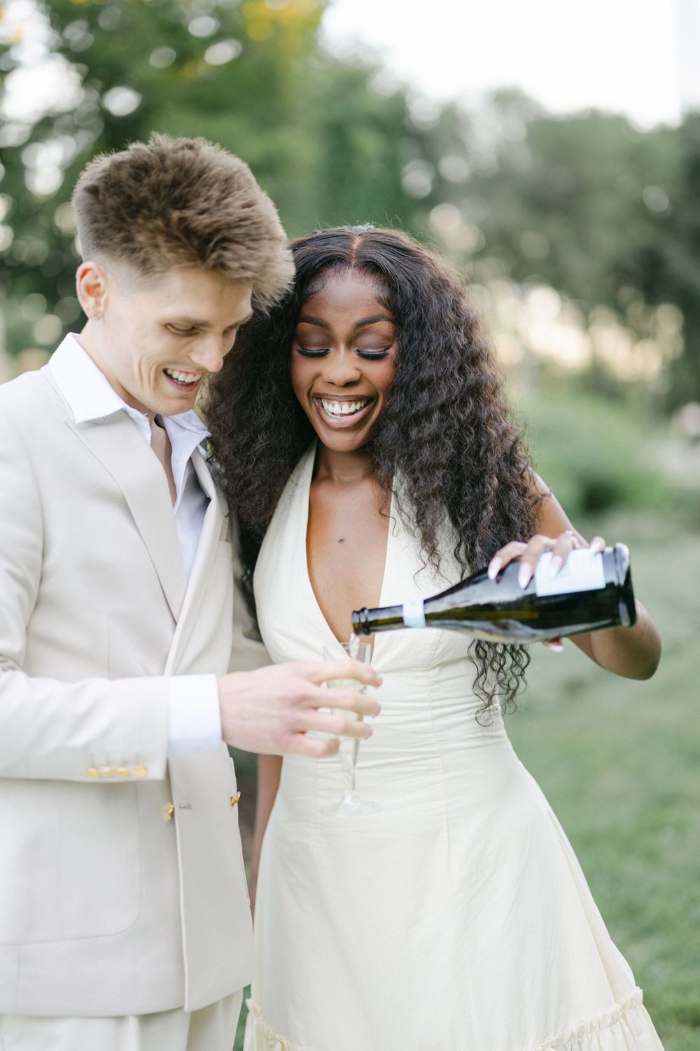 A joyful couple in cream-colored wedding attire share champagne together outdoors during a celebratory moment. - Grant Park Chicago