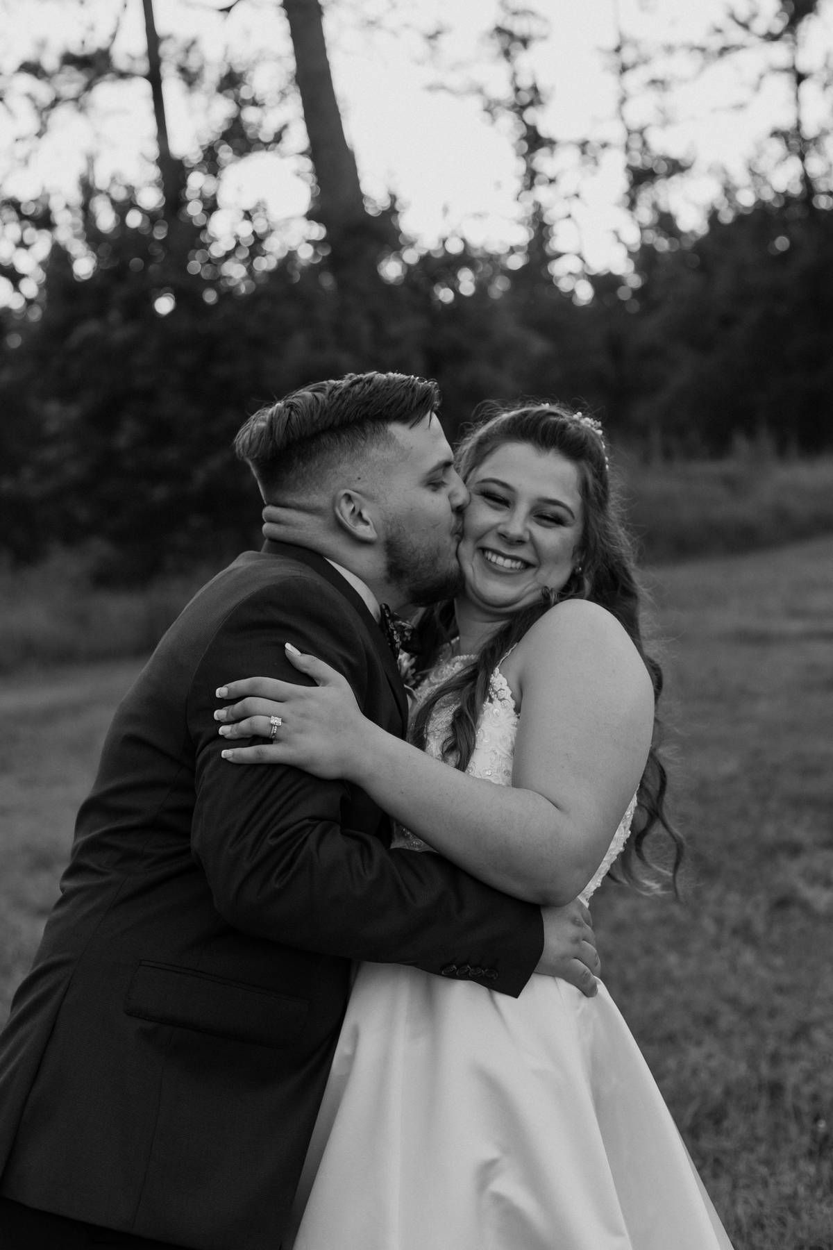 Black and white photo of a groom kissing the cheek of his smiling bride in a white gown while embracing outdoors in a field.