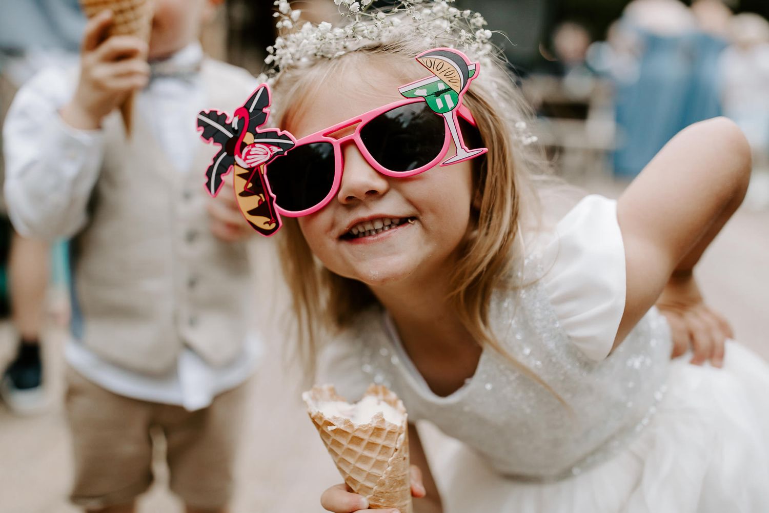 A joyful child in white wearing fun sunglasses and holding an ice cream cone at a summer celebration.