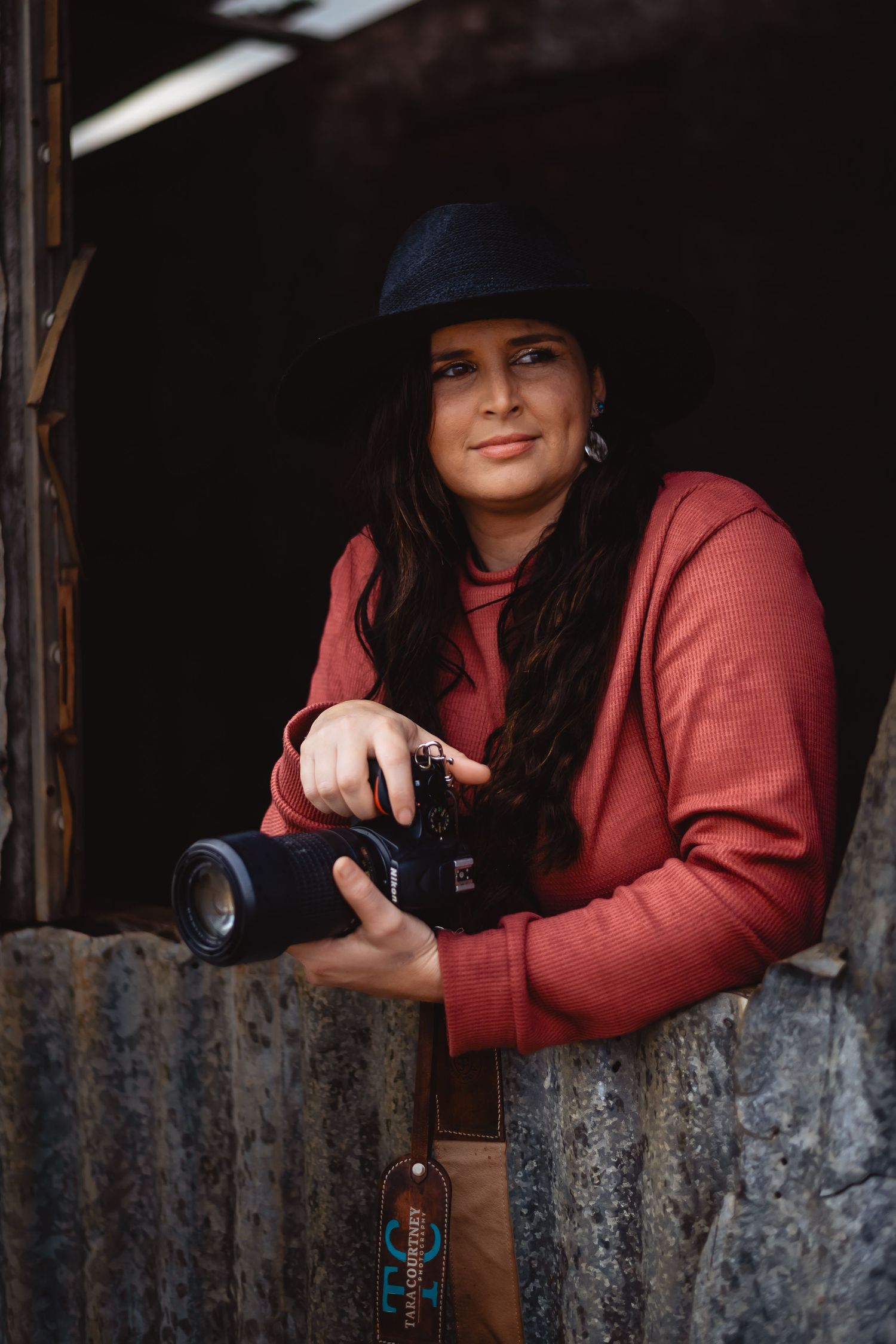 Photographer in red sweater and cap holds camera while standing in rustic barn doorway.