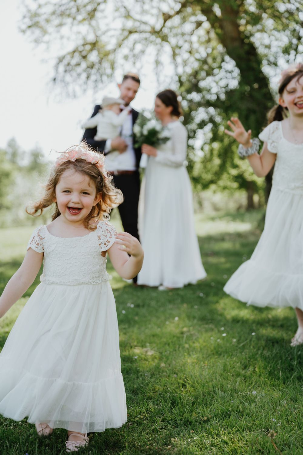 A flower girl in a white dress dances joyfully during an outdoor wedding celebration.