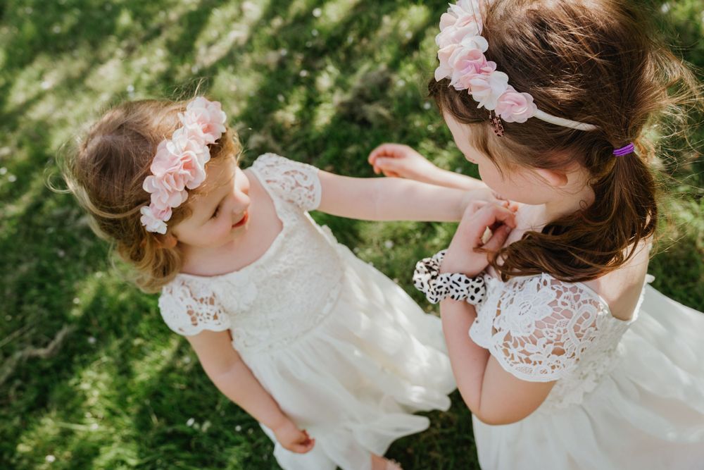 Two flower girls in white lace dresses share a playful moment outdoors.