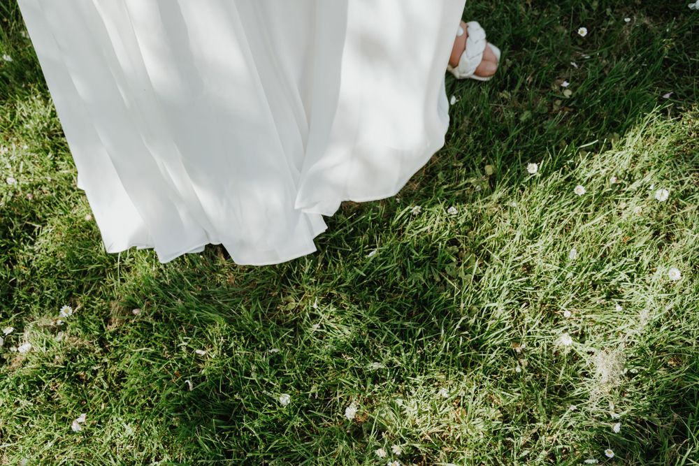 The hem of a flowing white wedding dress brushes against green grass in natural sunlight.