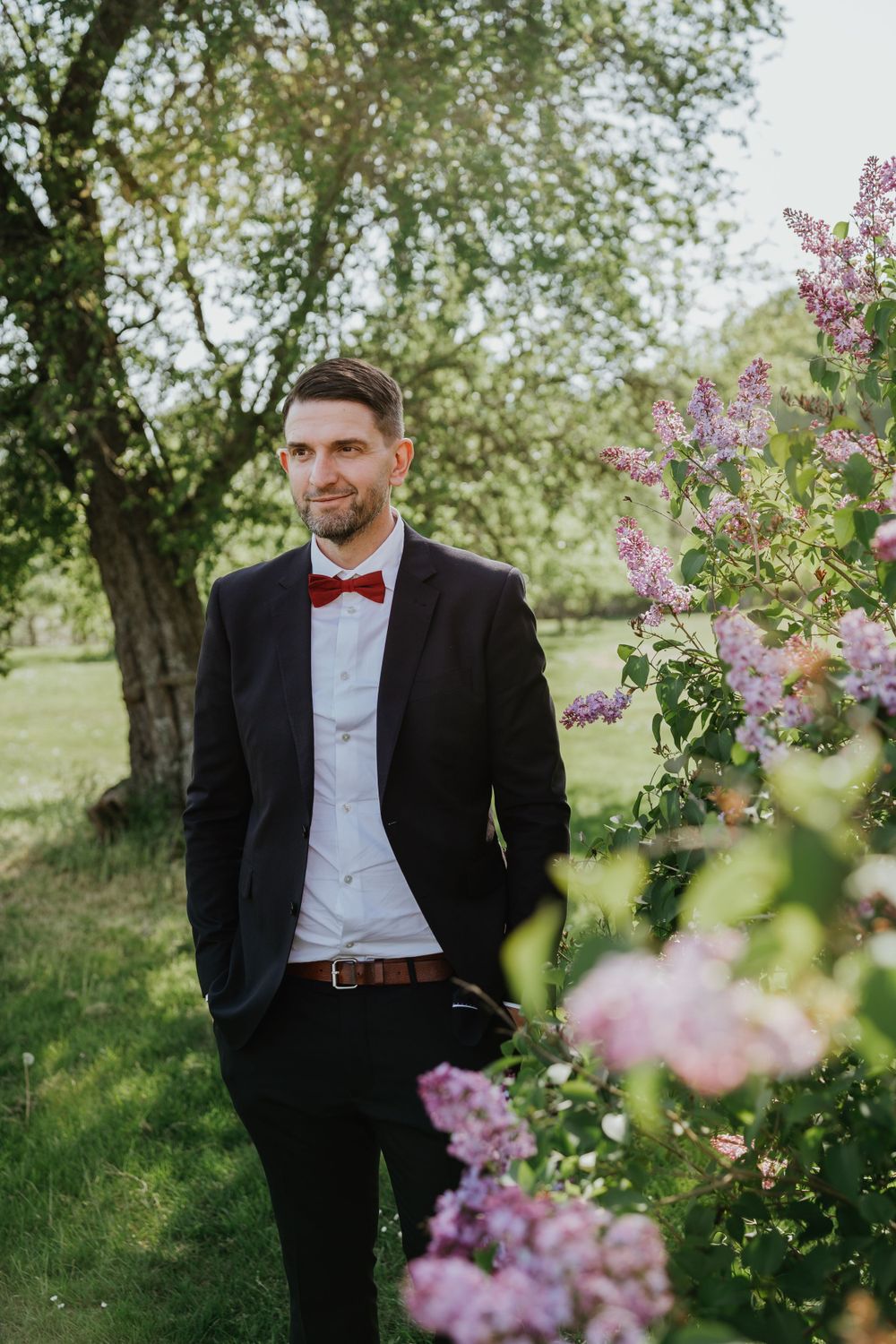 A formal portrait of a groom in a black suit with red bowtie among blooming lilacs.