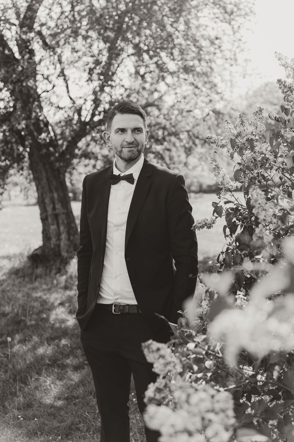 A black and white portrait of someone in a classic tuxedo and bowtie standing in a garden setting.