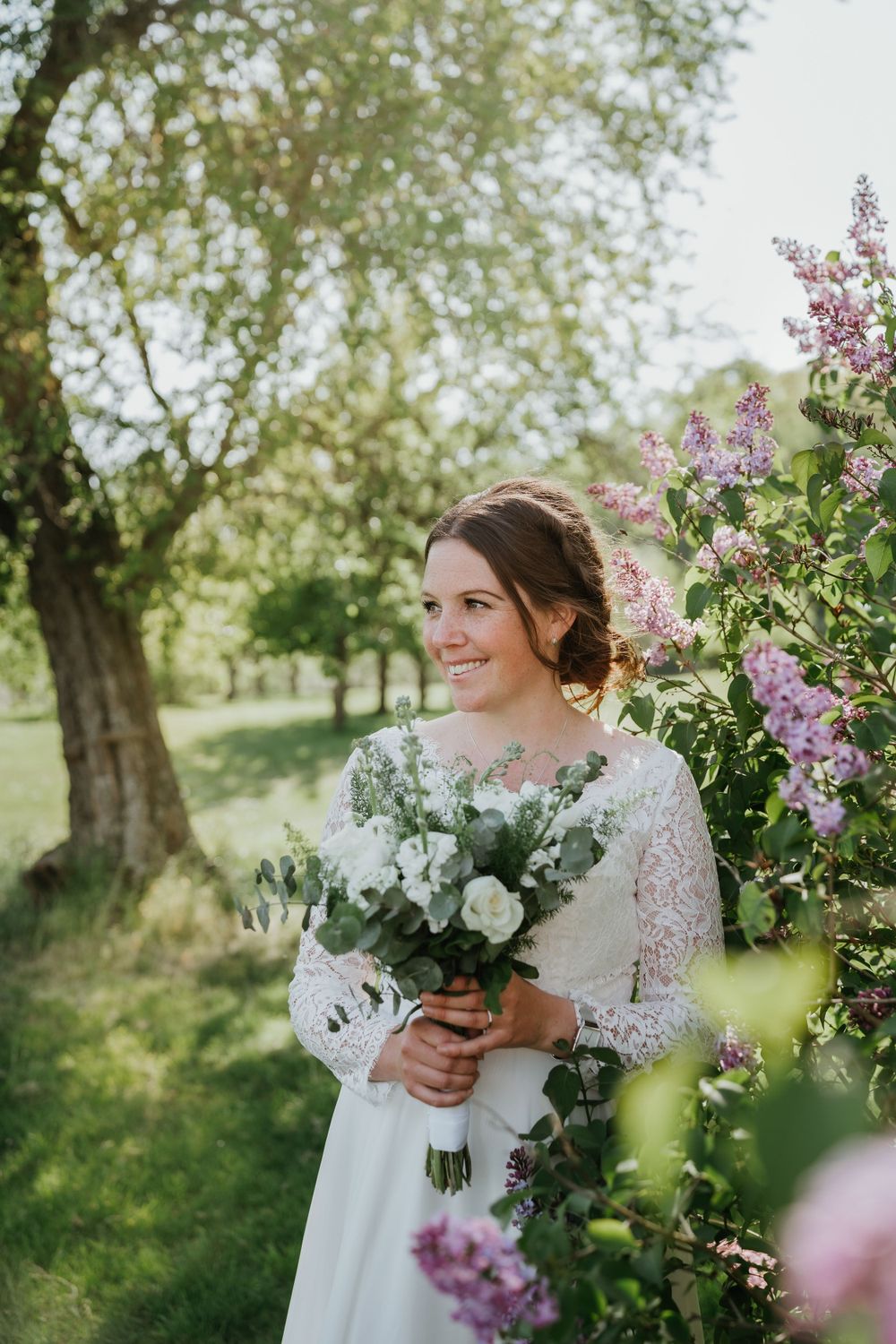 A bride in a lace sleeve wedding dress holds a white bouquet next to blooming lilac bushes.