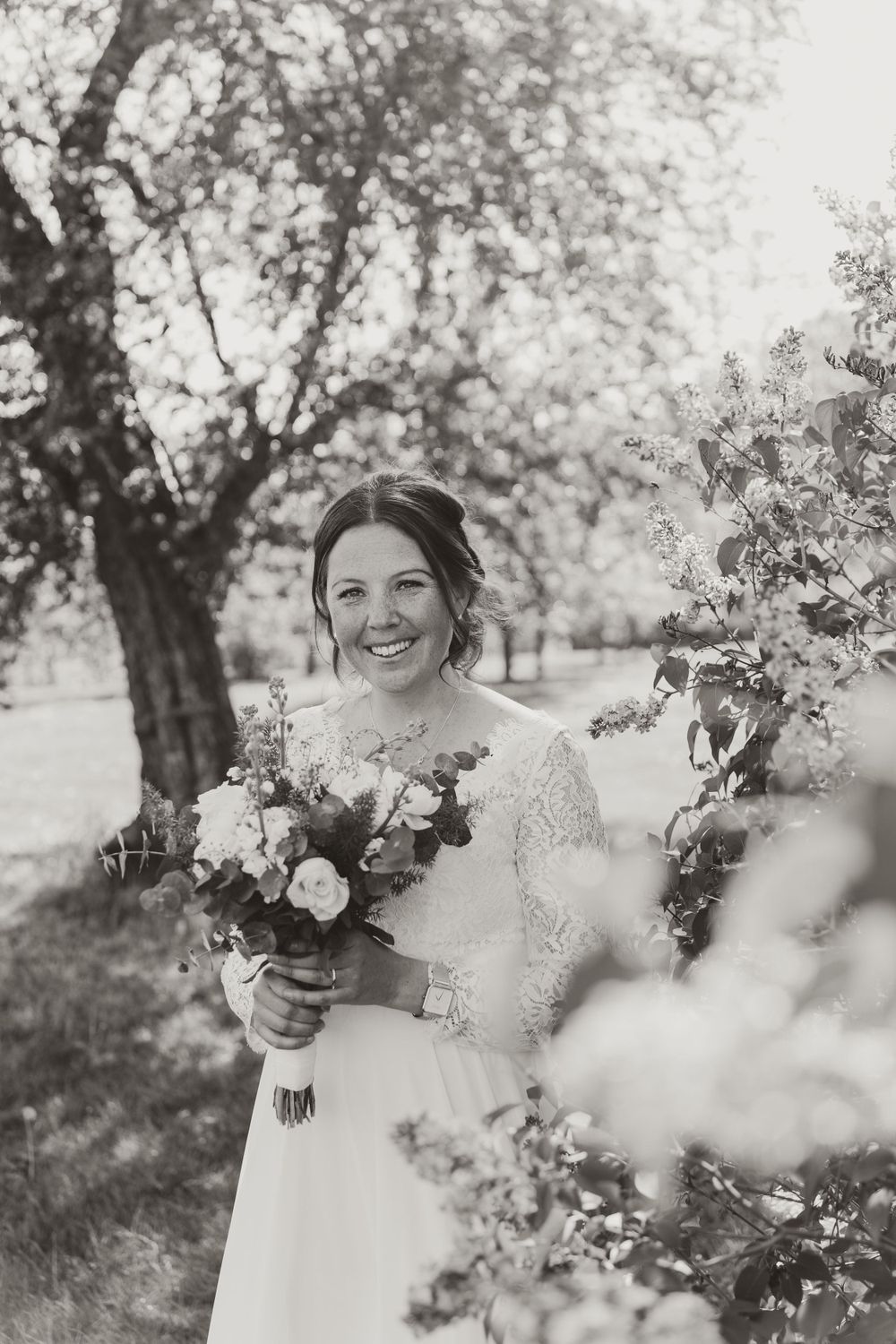 A black and white photo of a bride holding a bouquet and smiling outdoors among trees.