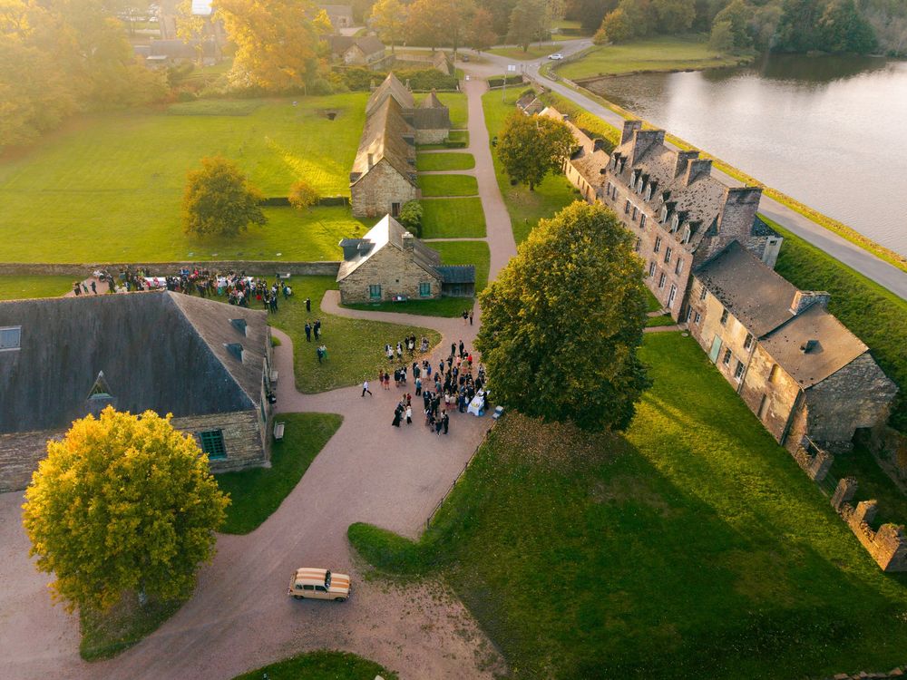 Aerial drone view of historic stone buildings along a waterfront with autumn trees and green fields in golden light.