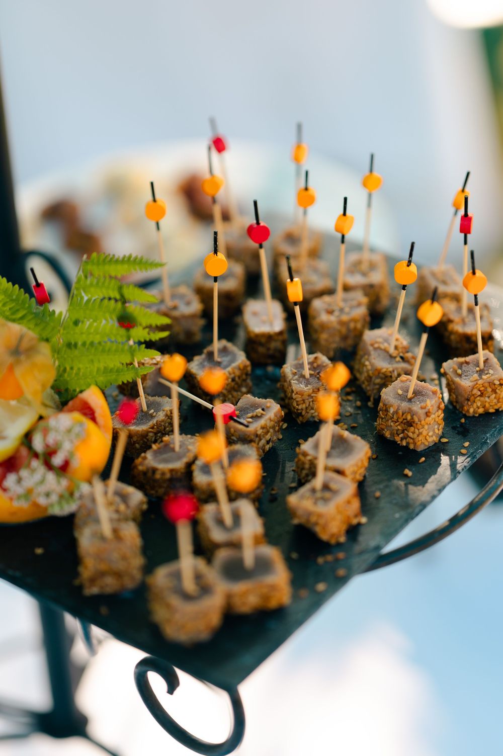 Appetizer bites topped with colorful garnishes and served on toothpicks displayed on a dark serving platter with ferns.