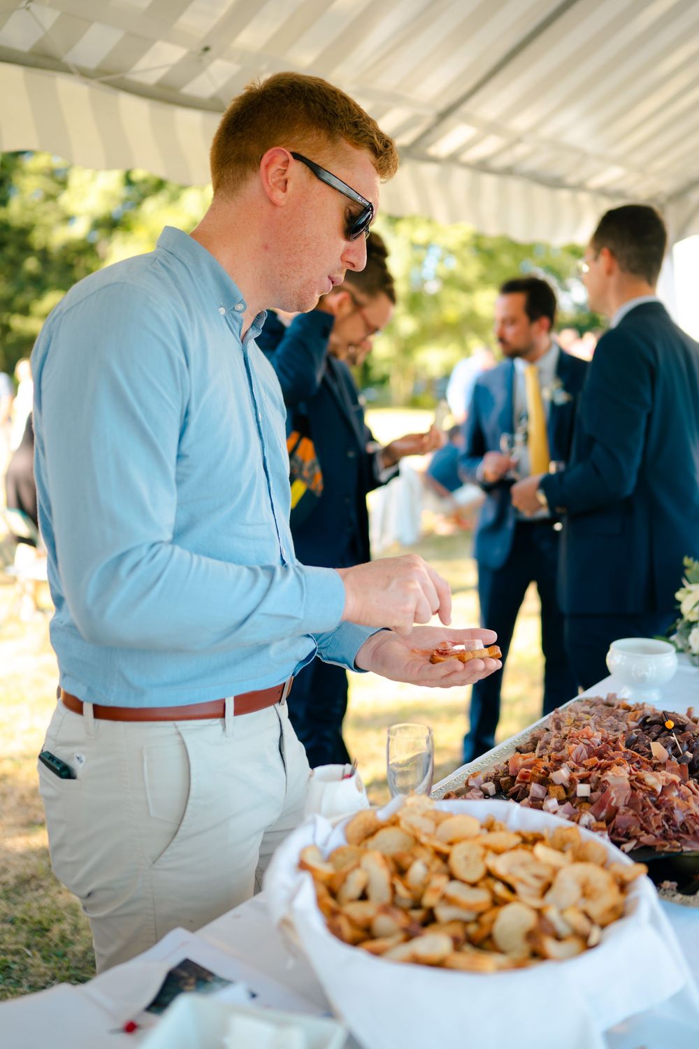 Guests in formal attire serve themselves at an outdoor buffet table with roasted meats and potatoes under a white tent.