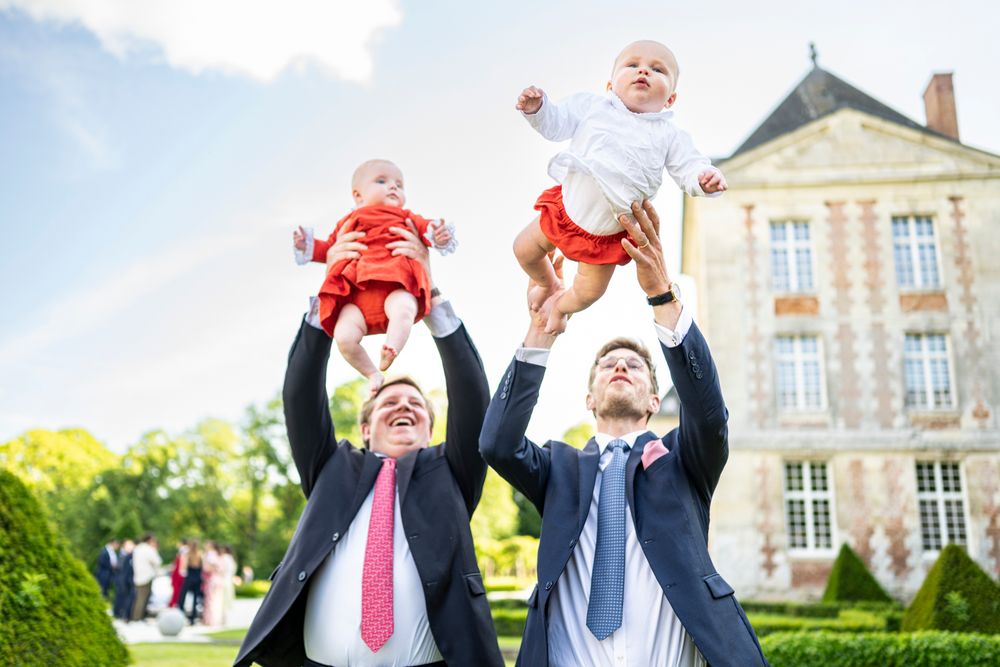 Two men in formal attire joyfully lift babies against a stately mansion backdrop.