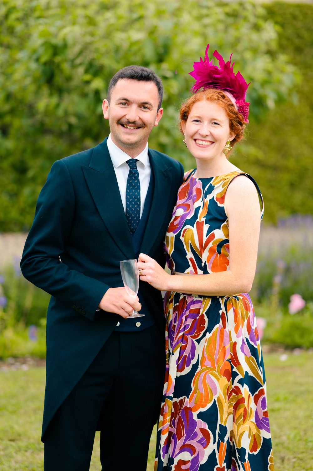 Elegant couple at outdoor event with vibrant floral dress and navy suit sharing drinks in garden setting.