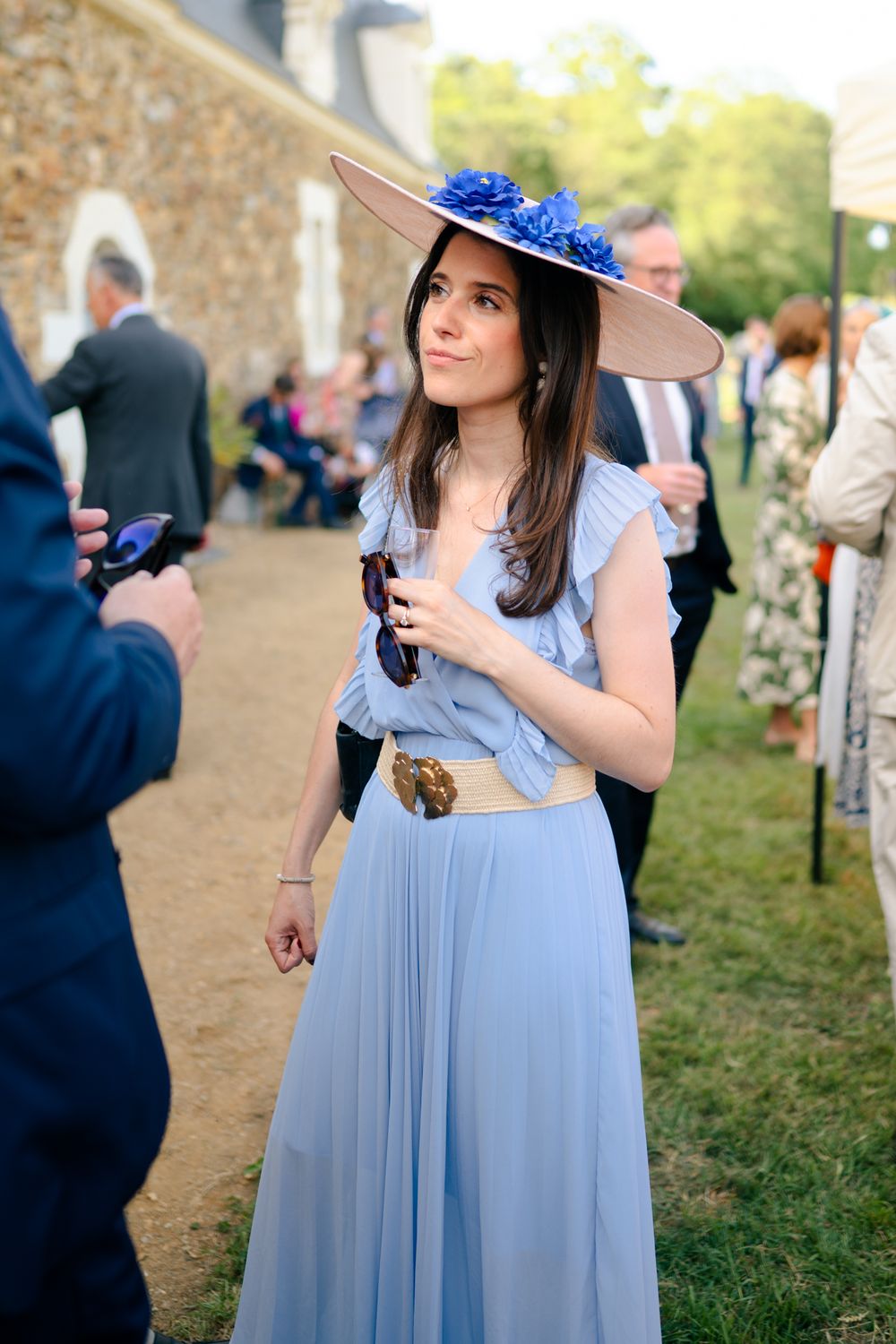 A person wearing a light blue dress and wide-brimmed hat attends an outdoor event in front of a stone wall.