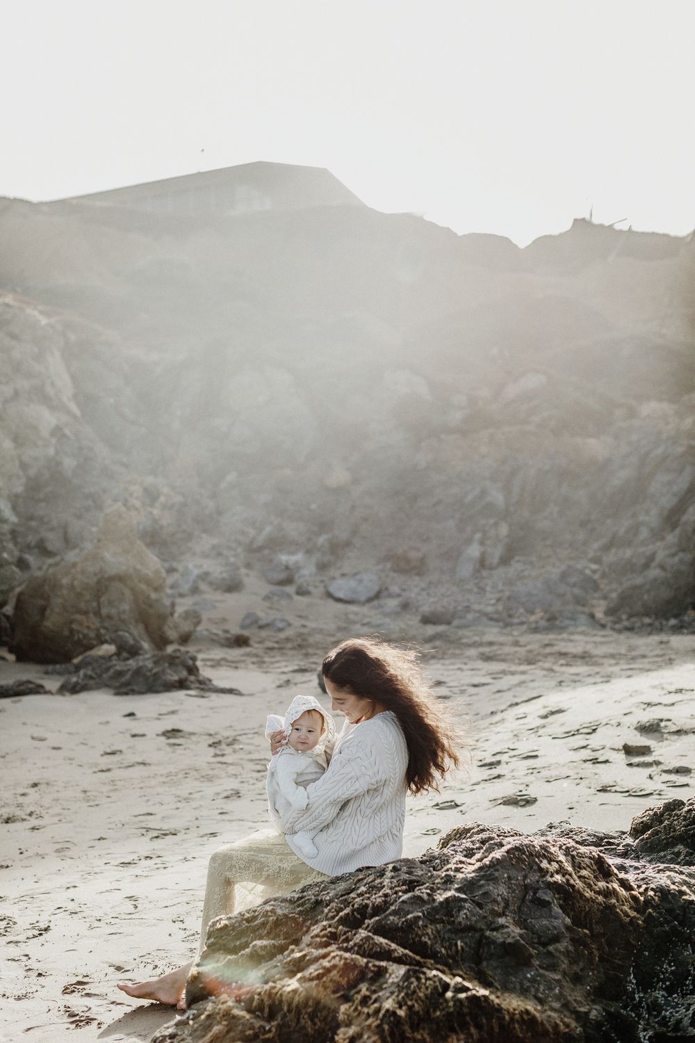 Mother with long hair sits on coastal rocks holds newborn baby with mist in golden sunlight background. North Shore motherhood photographer.