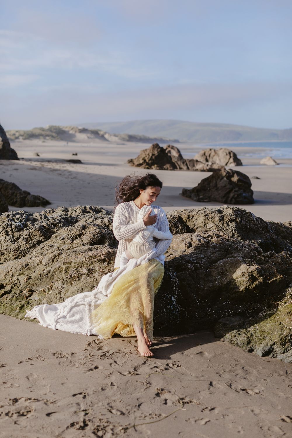 Mother with long dark hair holds newborn baby sitting on coastal rock with hills in background of ocean. North Shore family photographer