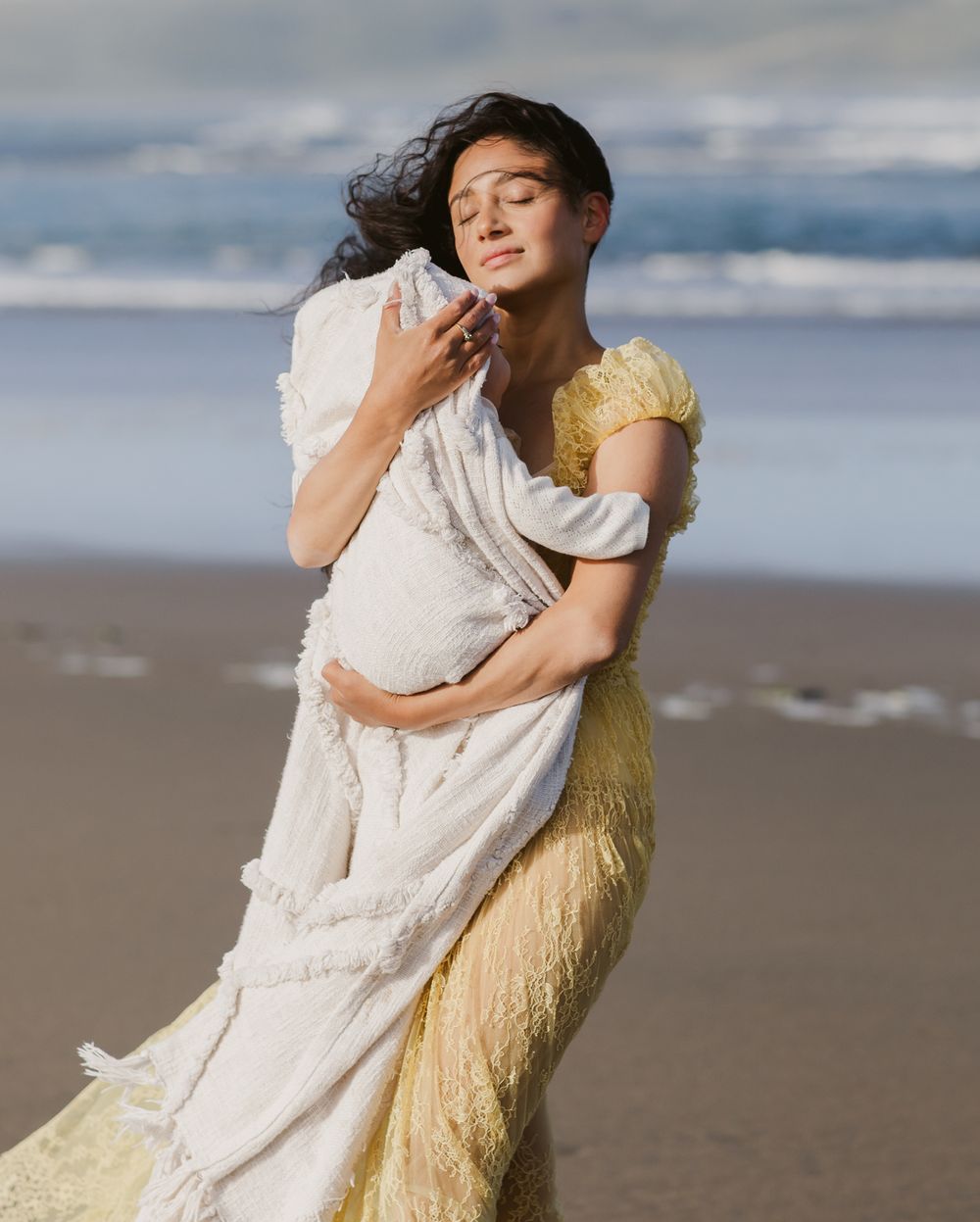 Mother with dark hair wearing yellow lace dress snuggles baby in blanket close at ocean. Kennebunkport Portsmouth family photographer