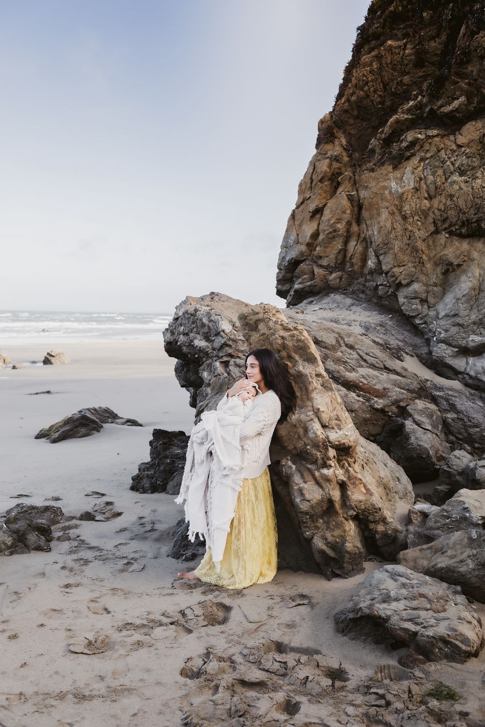 Mother with long hair snuggles baby in blanket leaning on large coastal rock. Lakes Region Southern NH motherhood photographer