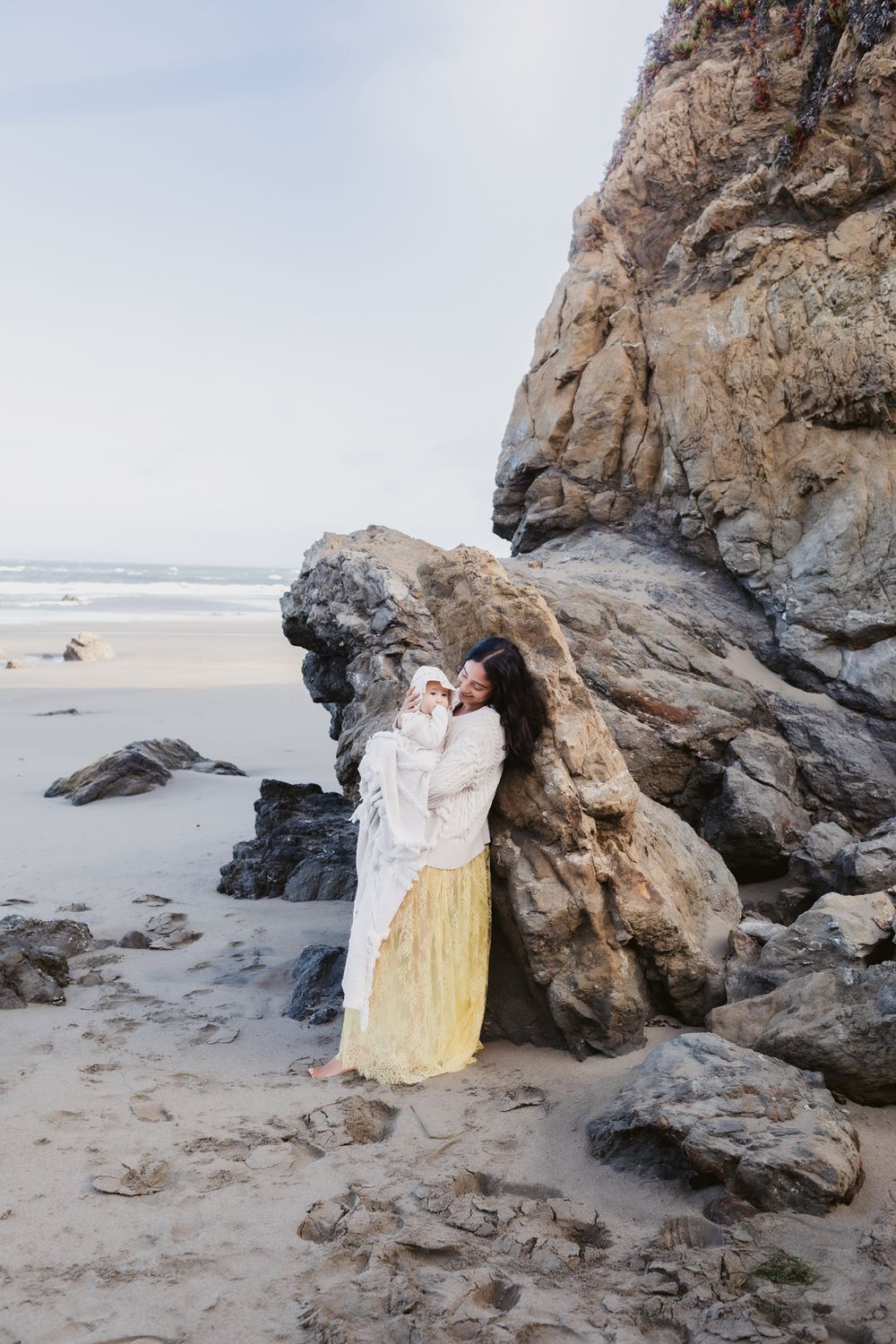 Mother with dark hair wears yellow lace dress holds baby in blanket leans against large coastal rocks at ocean. Hampton family photographer
