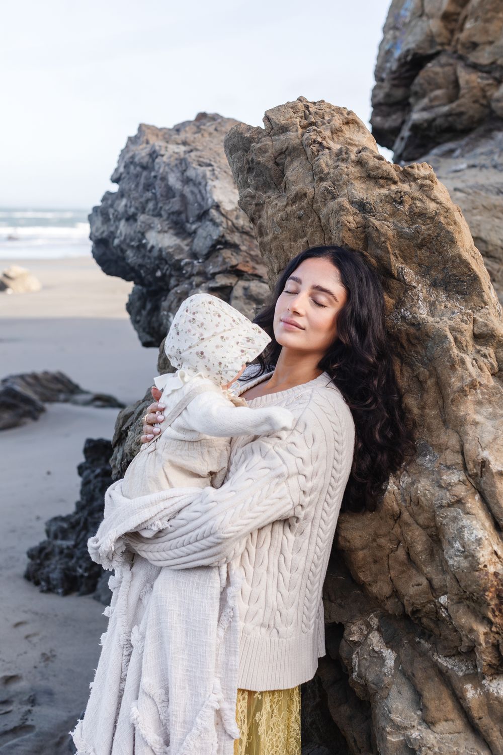 Mother with long hair holds baby in cream blanket leans on large rocks at beach. Gloucester, Concord, York, Acadia family photographer.