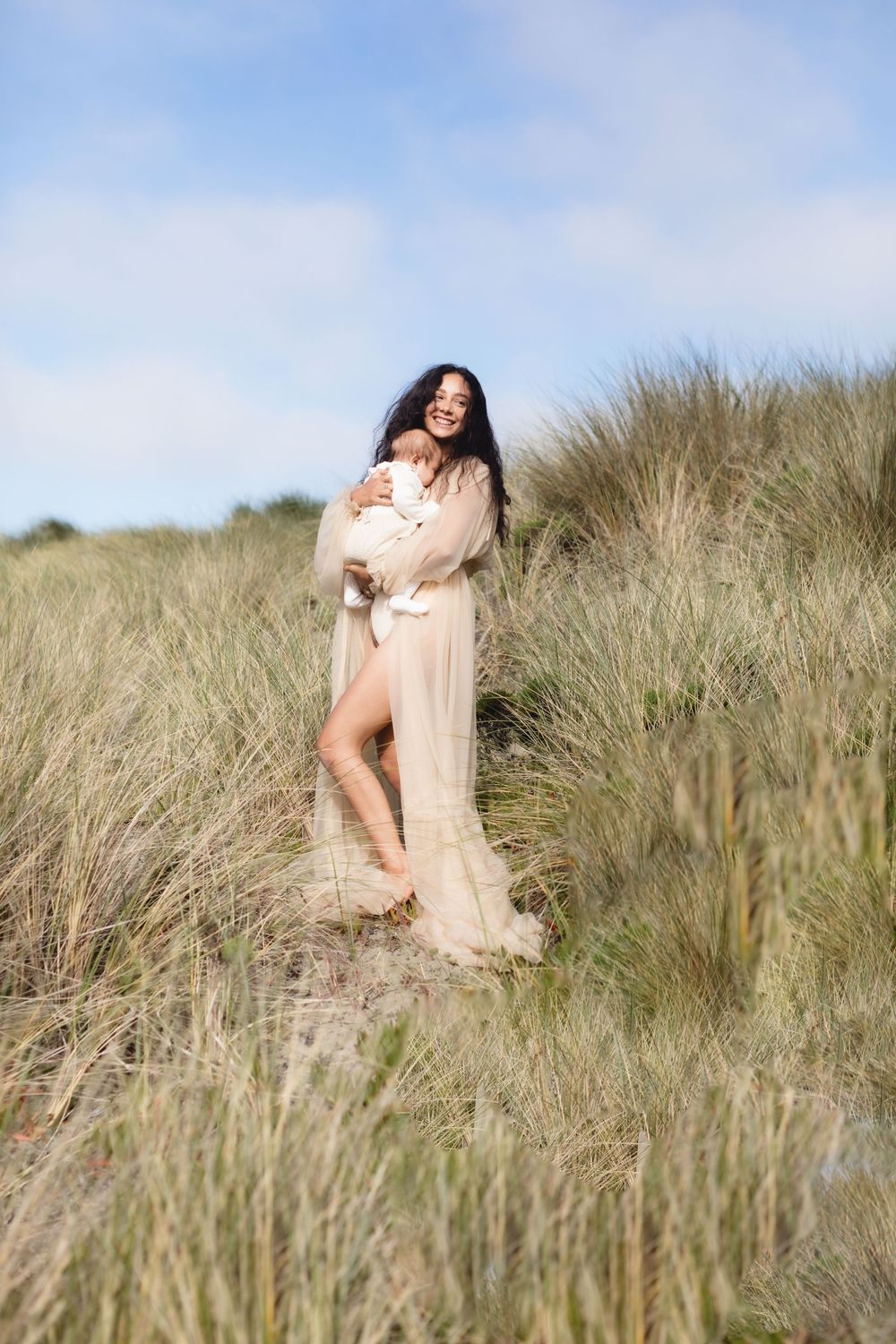 Mother wearing a long tan sheer dress laughs embracing her baby in coastal grass for motherhood photos. Southern Maine newborn photographer.