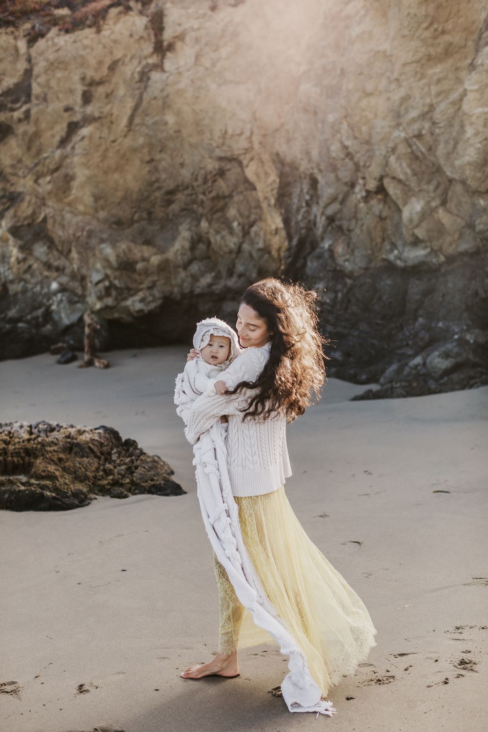 A mother in a yellow skirt and white sweater holds baby on sandy beach with large rock behind them. North Shore family photographer