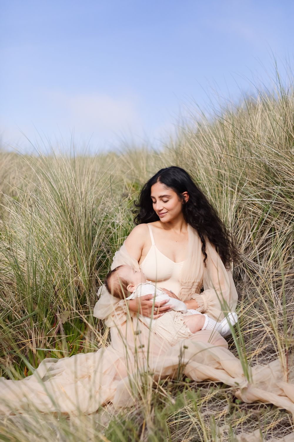 Mother with long dark hair wears a sheer tan covering holding baby girl in long seagrass on sunny day. Newburyport motherhood photographer