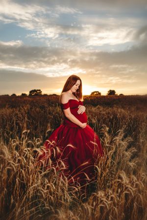 Woman in flowing red dress poses elegantly in wheat field during sunset. Middle TN Maternity Photographer