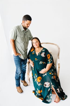 Couple in casual attire poses for maternity photos against white backdrop, woman wearing floral teal dress.