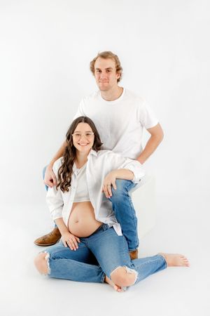 Couple in white shirts and jeans pose together on white background in casual maternity photoshoot.
